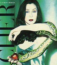 Woman seated with a green snake wrapped around her arms, holding an apple, with a CHER - IT'S A MANS WORLD (PREOWNED CD) poster in the background.