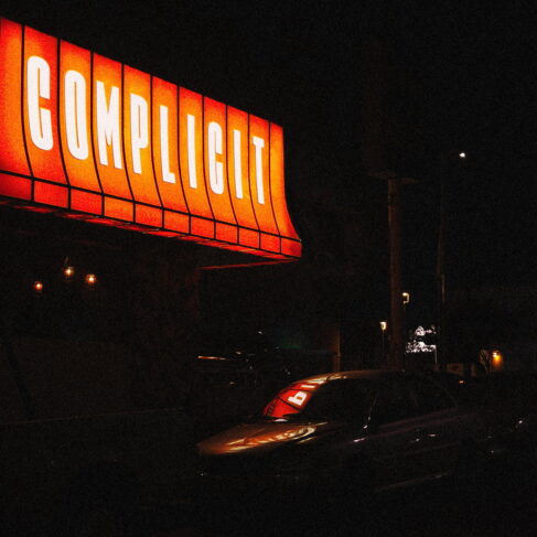 A neon sign that reads "ALEXISONFIRE - COMPLICIT (7" VINYL)" illuminates the night above a street scene on Vinyl Gold Coast, with a parked car under a muted streetlight.